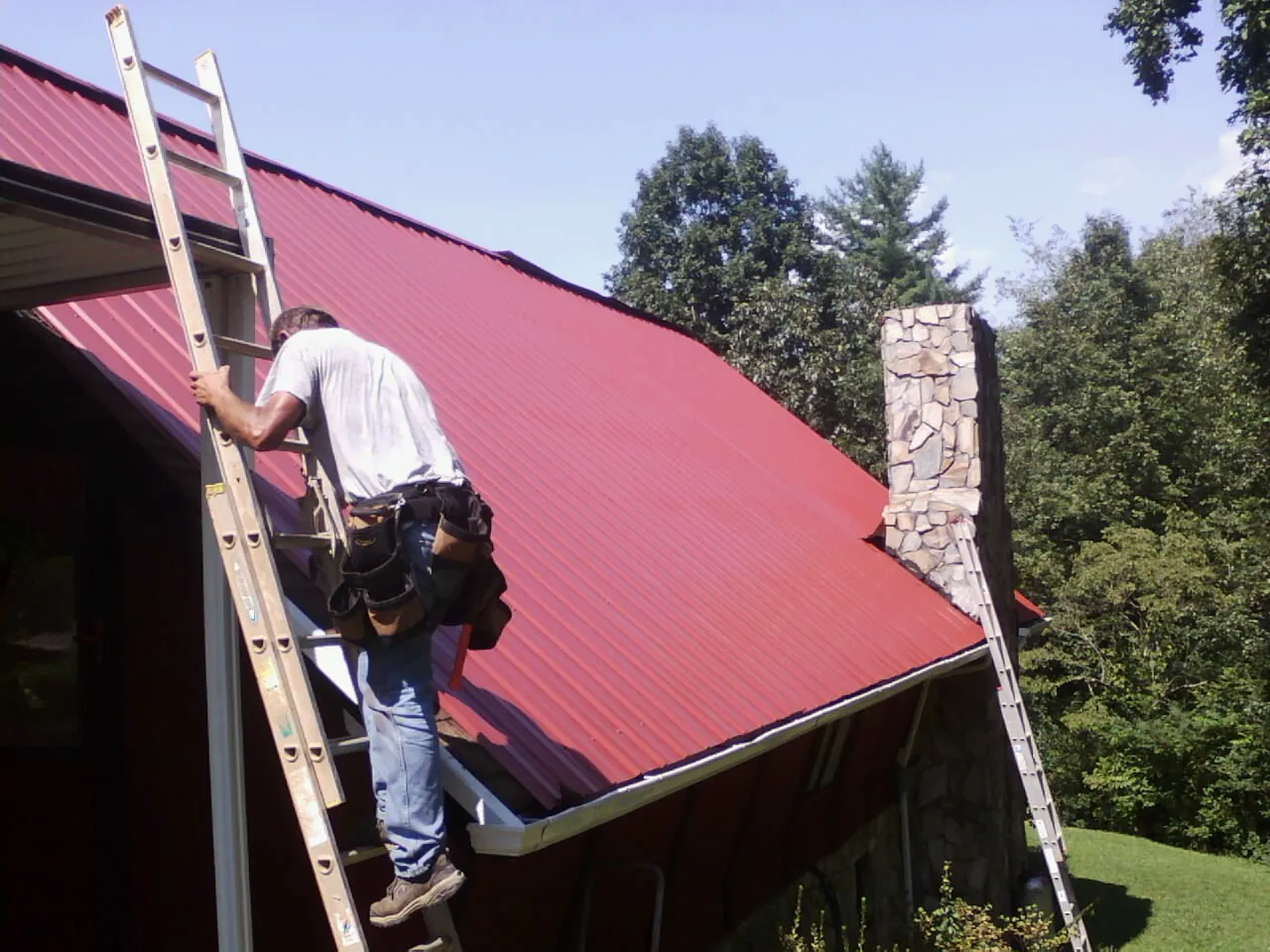 Roofer on a ladder beside a red metal roof and stone chimney — Soffit & Fascia Repair by Roofing Brothers in Estes Park